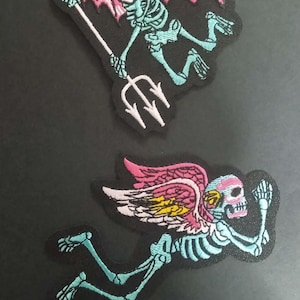 Peut inclure: Deux écussons brodés représentant des squelettes. L'un a des ailes de diable roses et un trident. L'autre a des ailes d'ange roses, jaunes et blanches. Les deux squelettes sont de couleur sarcelle avec des contours noirs.