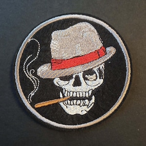 May include: Embroidered patch featuring a skull wearing a gray fedora with a red band, smoking a cigarette. The skull is white with black eye sockets and teeth, set against a black background with a silver border.