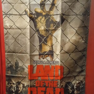 Puede incluir: Bandera rectangular con una mano de zombi que atraviesa una valla de alambre. La bandera tiene el texto "GEORGE A. ROMERO'S LAND OF THE DEAD" en naranja y blanco. La parte inferior muestra caras de zombis.