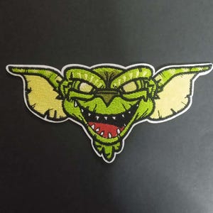 May include: Embroidered patch depicting a green creature with yellow ears, a wide grin, and red teeth. The patch has a white border, suitable for application on clothing or accessories.
