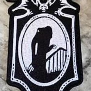 Peut inclure: Écusson brodé noir et blanc représentant une silhouette dans un cadre ovale. Le cadre est sur un fond noir avec une bordure décorative et un emblème en forme de chauve-souris en haut. L'écusson mesure environ 7.6 cm de haut.