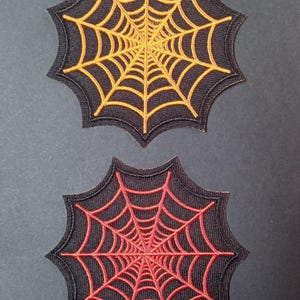 Puede incluir: Dos parches bordados con forma de telaraña. El parche superior presenta un fondo negro con un diseño de telaraña naranja, mientras que el parche inferior presenta un fondo negro con un diseño de telaraña roja. Ambos tienen bordes festoneados.