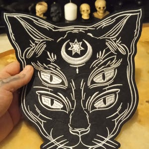 Peut inclure: Écusson de chat noir avec des détails brodés blancs. Le patch présente un visage de chat avec plusieurs yeux et un croissant de lune avec une étoile. L'écusson est un article sur le thème gothique ou occulte.