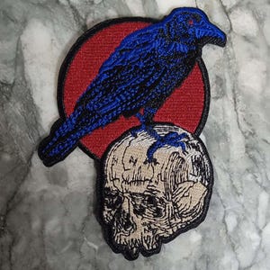 Peut inclure: Écusson brodé représentant un corbeau bleu aux yeux rouges perché sur un crâne. Le corbeau est posé sur un cercle rouge. Le crâne est détaillé avec des contours noirs et un remplissage beige, créant une esthétique gothique ou sombre.