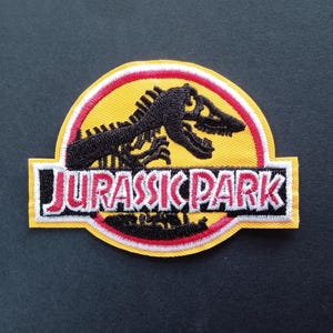 Peut inclure: Un écusson brodé représentant le logo de Jurassic Park. Le motif comprend la silhouette noire du squelette d'un Tyrannosaure Rex centrée sur un fond circulaire jaune avec une bordure rouge. Le texte JURASSIC PARK est en blanc et rouge.