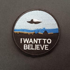 May include: A circular embroidered patch featuring a light blue sky, a dark blue mountain range, and a black foreground. A white UFO is centered in the sky. The text I WANT TO BELIEVE is embroidered in white capital letters across the black section.