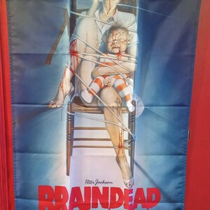 Peut inclure: Drapeau avec une illustration du film "Braindead" de Peter Jackson. L'image montre une femme en uniforme d'infirmière et un bébé, tous deux liés par des cordes et couverts de sang. Le titre du film est en rouge.