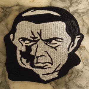 Peut inclure: Écusson brodé représentant un portrait en noir et blanc d'un homme au regard sévère. Le patch a une bordure noire et est destiné à être appliqué sur des vêtements ou des accessoires.