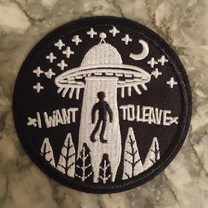 Könnte beinhalten: Schwarzer runder Aufnäher mit weißem Stickdesign. Das Design zeigt ein UFO, das eine Person hochbeamt, mit dem Text "I WANT TO LEAVE" sowie Sternen, einem Mond und Bäumen.