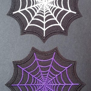 Peut inclure: Deux écussons brodés en forme de toiles d'araignée. L'un présente un motif de toile blanc sur fond noir, tandis que l'autre affiche un motif de toile violet sur fond noir. Les deux écussons ont un contour aux bords pointus bien définis.