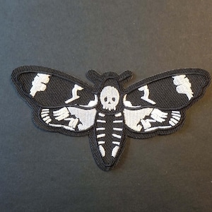 May include: Black and white embroidered moth patch. The patch features a moth design with a skull and skeleton details. The moth's wings and body are black, with white embroidered accents. The patch is likely for clothing or accessories.