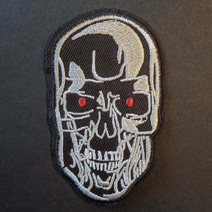 May include: Embroidered patch featuring a skull design with red eyes and silver detailing on a black background. The patch has a black border. This patch is suitable for jackets, bags, or other fabric items.