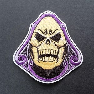 Peut inclure: Un écusson brodé représentant un personnage de crâne stylisé avec un visage jaune pâle, des orbites noires profondes et une mâchoire saillante. Le personnage est encadré par une capuche violette vive avec des motifs tourbillonnants et un bord blanc.