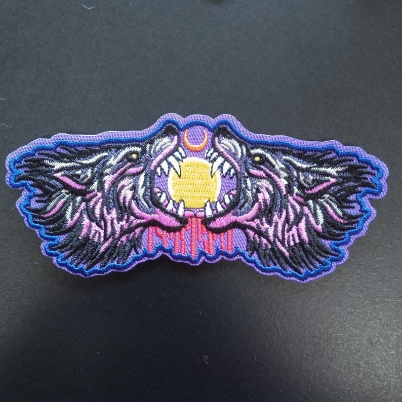 K&ouml;nnte beinhalten: Ein gestickter Aufn&auml;her mit zwei knurrenden Wolfsk&ouml;pfen, die sich gegen&uuml;berstehen, mit einer gelben Sonne und einem kleinen orangefarbenen Halbmond dazwischen. Das Design verwendet schwarzes, violettes, rosa und gelbes Garn mit einem violetten Rand.