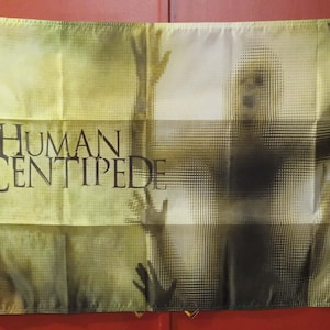 Puede incluir: Una bandera con el texto "THE HUMAN CENTIPEDE" en letras negras. La imagen muestra una figura pixelada con los brazos levantados. El fondo es de color verde descolorido.