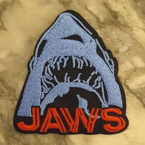 Op de afbeelding: Geborduurde patch met een open haaienbek met tanden. De haai is lichtblauw, tegen een zwarte achtergrond, met het woord "JAWS" in het rood. De patch heeft een zwarte rand.