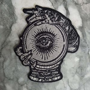 Peut inclure: Écusson brodé représentant une boule de cristal avec un motif d'œil. L'écusson est dans les tons de gris et de noir, avec des détails complexes. Une main tient la boule de cristal. L'écusson a une bordure décorative.