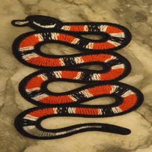 May include: Embroidered snake patch in a coiled design. The patch features a black outline with alternating bands of red, white, and black. The snake's head is at the top, with a white and black pattern.