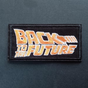 Peut inclure: Un écusson rectangulaire brodé sur fond noir avec le texte BACK TO THE FUTURE en lettres dégradées orange et jaune. Le design comprend un graphique en forme de flèche stylisée s'étendant sur le côté droit du mot BACK.