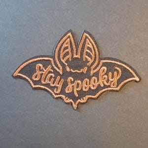 Könnte beinhalten: Schwarzer fledermausförmiger Aufnäher mit dem Schriftzug "Stay Spooky" in Kupfer gestickt. Der Aufnäher zeigt ein detailliertes Fledermausdesign mit ausgebreiteten Flügeln, ideal für Halloween.