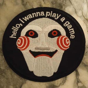 May include: A circular black patch with a white puppet face, red spiral cheeks and eyes. The text "hello, I wanna play a game" is embroidered in white around the edge.