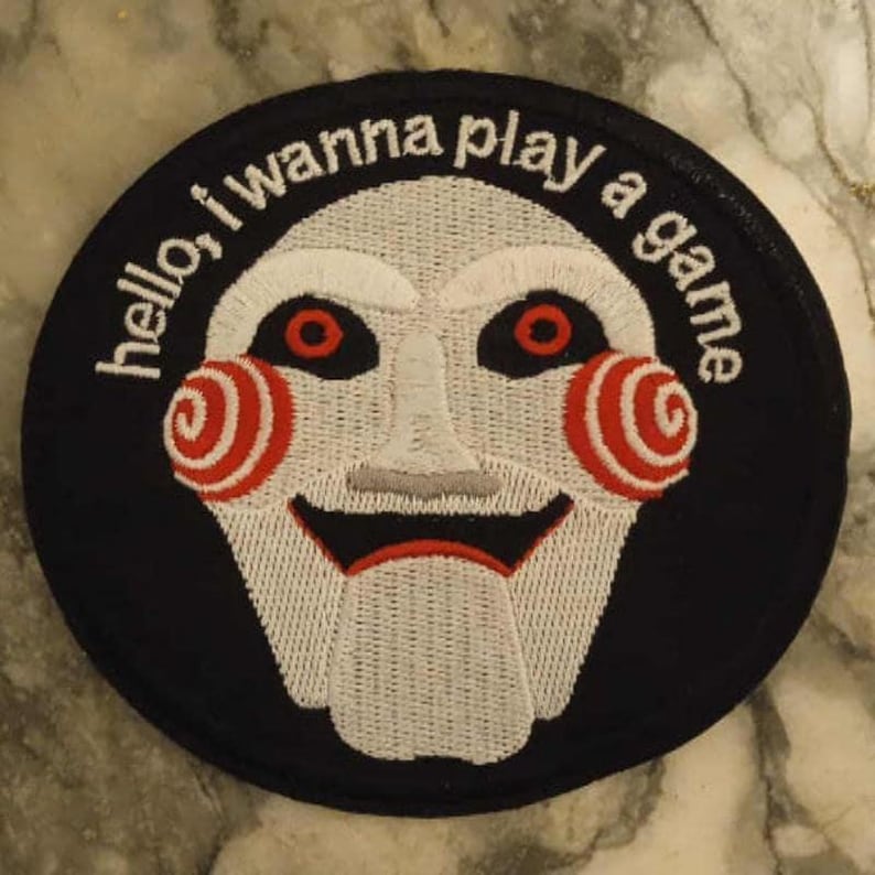 May include: A circular black patch featuring a white puppet face with red spiral cheeks and eyes. The text "hello, I wanna play a game" is embroidered around the edge in white.