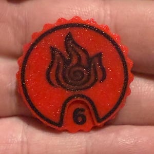 May include: A red, circular game piece with a black flame design and the number 6. The piece has a textured, glittery surface and a black border. The flame design is in the center.