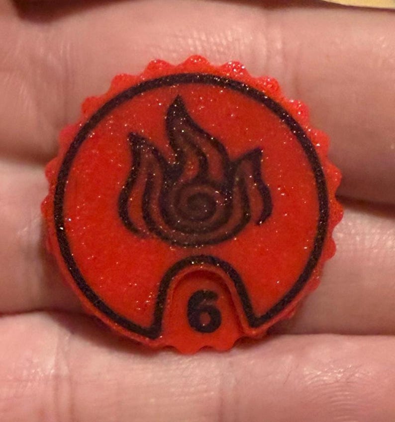 May include: A red, circular game piece with a black flame design and the number 6. The piece has a textured, glittery surface and a black border. The flame design is in the center.