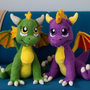 May include: Two crocheted dragon toys. One is green with yellow and orange accents, the other is purple with yellow and orange accents. Both have white horns and eyes, and are sitting on a blue surface.