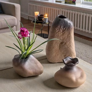 May include: Three ceramic vases in varying shapes and sizes sit on a light-colored wooden table. The vases have a stone-like appearance with a gradient of brown and gray tones. One vase holds a plant with green leaves and a pink flower.