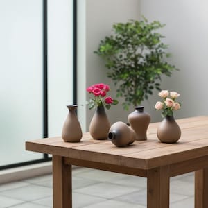 May include: A selection of small, brown ceramic vases with dark grey rims, some holding pink and peach roses. The vases are displayed on a wooden table, creating a decorative arrangement. A green plant is in the background.