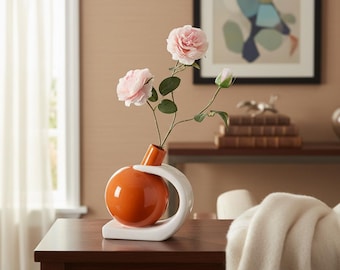 Pode incluir: Um vaso esférico laranja brilhante com base curva e suporte de pescoço brancos, contendo duas rosas artificiais rosa claro e um botão fechado num caule verde fino. O vaso apresenta uma superfície lisa, reflexiva e um design escultural.