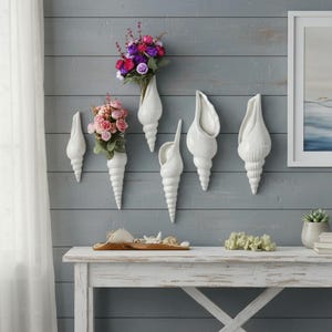May include: White ceramic seashell wall vases, some filled with colorful flowers, are mounted on a gray-blue wooden wall. The vases vary in size and shape, creating a coastal, decorative theme. A white table below displays seashells and decor.