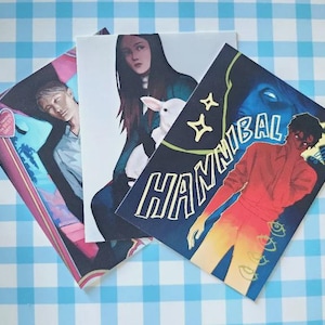 Puede incluir: Una colección de grabados artísticos con ilustraciones de personas y la palabra "HANNIBAL" en blanco. Los grabados tienen una variedad de combinaciones de colores, incluyendo rosa, azul y rojo, y están dispuestos sobre una superficie a cuadros azul y blanco.