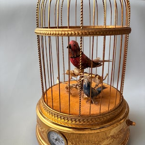 May include: A gold-colored decorative birdcage with a clock and two small birds inside. The cage has a domed top and a round base with ornate detailing. The birds are perched on a branch inside the cage.