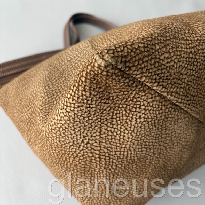 May include: Close-up of a brown tote bag with a textured, dotted pattern. The bag has brown handles and a structured shape. The material appears to be leather or a leather-like fabric.