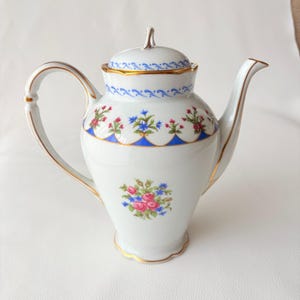 May include: A white porcelain teapot featuring a floral pattern of pink roses and blue cornflowers. The teapot has a gold-trimmed handle, spout, and lid, with a blue scalloped border design around the upper section and a central floral bouquet.
