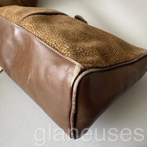 May include: A brown leather handbag with a textured, patterned top section. The bag features a rectangular shape with rounded corners and a visible buckle. The leather appears worn, with visible stitching and a contrasting texture.