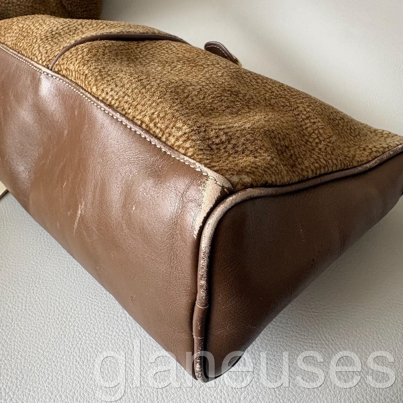 May include: A brown leather handbag with a textured, patterned top section. The bag features a rectangular shape with rounded corners and a visible buckle. The leather appears worn, with visible stitching and a contrasting texture.