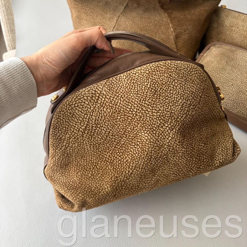 May include: A brown and tan handbag with a textured exterior and a rounded shape. The bag has a dark brown handle and trim, with gold-colored hardware accents. The bag is held by a person.