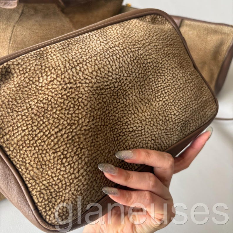 May include: A set of brown leather bags with a textured surface. The bags have a rectangular shape with rounded corners and a darker brown trim. The bags are held in a person's hand.