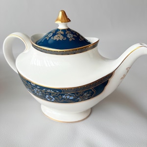 Puede incluir: Una tetera de porcelana blanca con una banda azul oscuro decorada con intrincados motivos florales en azul claro y bordes dorados. La tapa tiene un remate dorado, y el cuerpo incluye un asa y un pico curvos con detalles dorados a juego.
