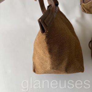 May include: A brown handbag with a textured exterior and a leather handle. The bag has a structured shape and a contrasting brown leather trim. The bag is held up against a white background.