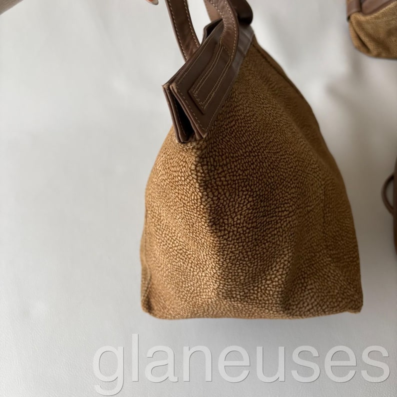 May include: A brown handbag with a textured exterior and a leather handle. The bag has a structured shape and a contrasting brown leather trim. The bag is held up against a white background.