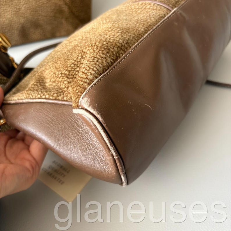 May include: A brown handbag with a textured, speckled upper section and smooth, dark brown leather sides. The bag has a rectangular shape with a white trim and a long strap. A tag with text is attached.