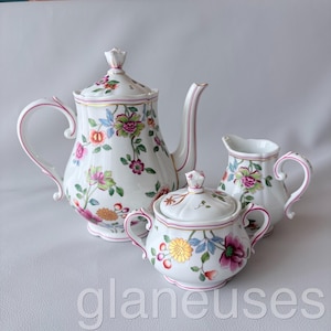 Op de afbeelding: Een wit porseleinen theeservies met een theepot, melkkan en suikerpot, allemaal versierd met een kleurrijk bloemmotief. De set heeft roze accenten en gouden randen. De theepot heeft een deksel met een decoratieve knop.