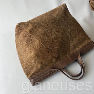 May include: A brown tote bag with a textured exterior and leather handles. The bag features a structured design with a rectangular base and a spacious interior. The handles are a darker shade of brown, and the bag has a stitched trim.