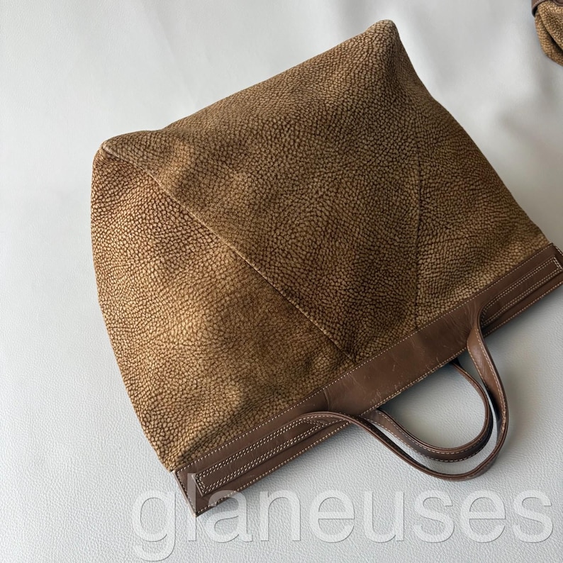 May include: A brown tote bag with a textured exterior and leather handles. The bag features a structured design with a rectangular base and a spacious interior. The handles are a darker shade of brown, and the bag has a stitched trim.