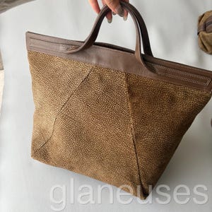 May include: A brown tote bag with a textured exterior and brown leather handles and trim. The bag has a rectangular shape with a wide opening and a stitched leather border. The bag is held by a person.