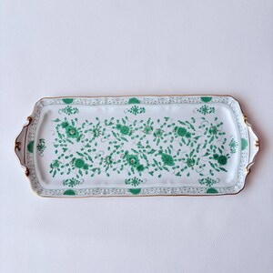 以下が含まれることがあります： 白いベースに緑の花柄が描かれた長方形の磁器製サービングトレイ。トレイの縁はスカラップ状で金色の縁取りが施され、両側に装飾的なハンドルが付いています。中央には繊細な緑の蔓と花が描かれています。